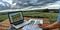 Person planning vineyard layout with a laptop, calculator, and vineyard row diagrams on a wooden table overlooking farmland under cloudy skies.